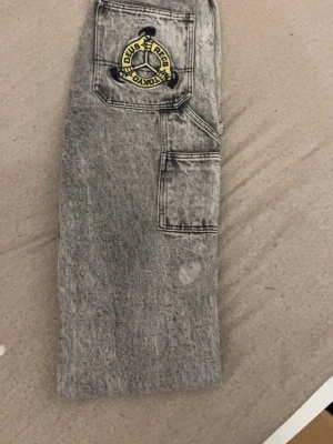 Grå cargobyxor med patch - Säljer ett par grå cargobyxor i tvättad denim-look med flera fickor och en stor patch med texten 'DEUS EX MACHINA TOKYO' på ena benet. Byxorna har raka ben och tydliga sömmar för en cool streetwear-stil. Perfekta för dig som gillar utility och trendiga detaljer.