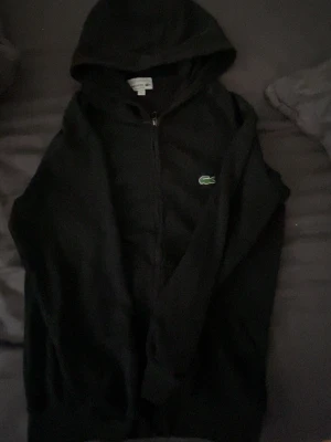 Svart zip hoodie från Lacoste - Säljer en svart hoodie från Lacoste med dragkedja och huva. Klassisk krokodil-logga på bröstet och regular fit. Perfekt för dig som gillar stilrena och bekväma plagg. Materialet känns mjukt och skönt mot huden.