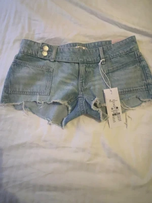 Ljusblå jeansshorts med rå kant - Säljer ett par ljusblå jeansshorts med fransig, rå kant och stora fickor både fram och bak. Shortsen har dubbla silverknappar i midjan och bälteshällor. Perfekta för sommaren och riktigt snygga till en avslappnad stil.  Säljer dessa shorts ifrån gina 14 + , lappen är kvar då jag glömde skicka tillbaka dom i tid och dom inte passade. Ny pris 299