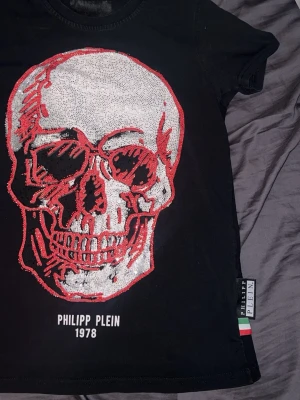 Svart Philipp Plein t-shirt med dödskalle - Snygg svart t-shirt från Philipp Plein med en stor glittrig dödskalle i silver och rött framtill. T-shirten har korta ärmar och rund hals. Märkeslapp på sidan med italienska flaggan. Perfekt för dig som gillar statement-plagg.