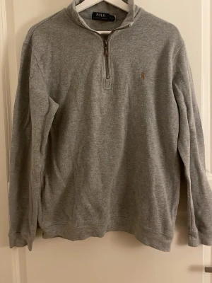 Grå half zip tröja från Polo Ralph Lauren - Klassisk grå långärmad tröja från Polo Ralph Lauren med half zip och liten broderad logga på bröstet. Tröjan har en hög krage och är tillverkad i mjukt bomullsmaterial. Perfekt för en avslappnad och stilren look.
