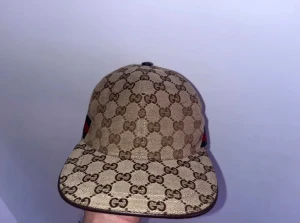 Beige keps från Gucci med GG-mönster - Säljer en beige keps från Gucci med klassiskt GG-monogram i brunt över hela kepsen. Kepsen har böjd skärm och en grön-röd rand på sidan. Tillverkad i canvasmaterial med detaljer i svart. Perfekt för dig som gillar exklusiv streetwear.