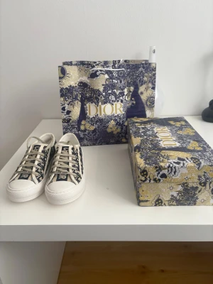 Dior sneakers i blått och vitt - Snygga Dior sneakers i lågt snitt med klassiskt blått och vitt mönster och tydlig Christian Dior-logga på sidan och tungan. Skorna har vita snören och sula i gummi. Perfekta för dig som vill ha en exklusiv och trendig look.
