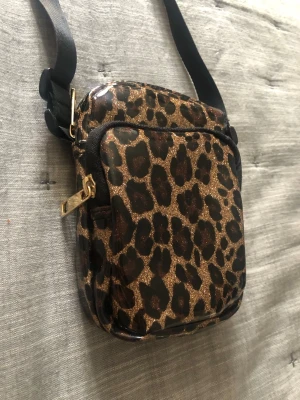 Leopardmönstrad axelväska med glitter - Snygg axelväska i leopardmönster med glittrig finish. Väskan har justerbar svart axelrem, guldfärgade dragkedjor och två fack. Perfekt storlek för mobilen och småprylar, ca 16,5 x 10 cm. Materialet är glansigt och lätt att torka av.