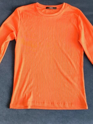 Ribbad orange långärmad topp - Snygg ribbad topp i stark orange färg med lång ärm och rund hals. Tillverkad i ett stretchigt material som ger en bekväm passform. Perfekt för dig som vill sticka ut med färg och enkel stil. Storlek! Axelbredd 42cm och längd 60cm