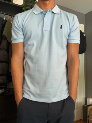 Ralph Lauren Piké – Ljusblå - Säljer en klassisk Ralph Lauren piké i en stilren ljusblå färg med mörkblå broderad logga på bröstet. En tidlös modell som passar lika bra till vardags som till golf eller en mer uppklädd look. Storleken är medium och sitter true to size. Skicket är mycket bra då den aldrig använts