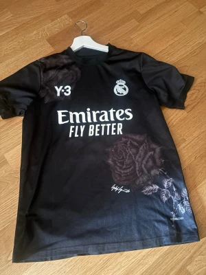 Real Madrid svart matchtröja Y-3 - Snygg svart Real Madrid matchtröja i samarbete med Y-3 och Adidas. Tröjan har vita tryck med klubbmärke, sponsor och nummer 22 (Rüdiger) på ryggen. Unika rosdetaljer på både fram- och baksida samt signaturtryck. Tillverkad i lätt och funktionellt material.