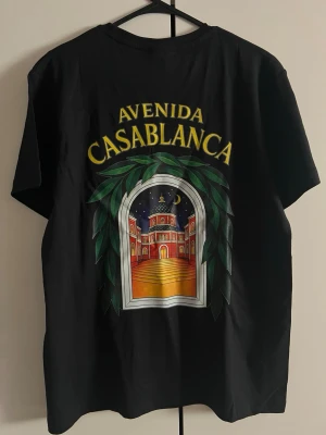 Svart t-shirt Avenida Casablanca - Svart t-shirt med tryck både fram och bak. På ryggen står det 'Avenida Casablanca' i gult ovanför ett färgglatt motiv av en byggnad i nattmiljö, inramad av gröna blad. Framtill finns en mindre version av samma tryck. Rund hals och korta ärmar.