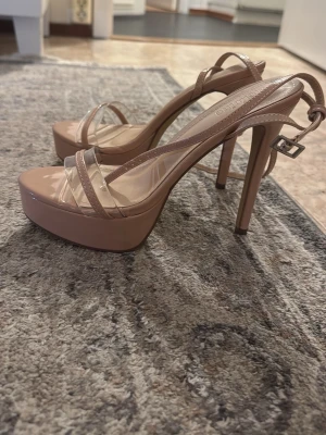 Nude platåpumps med transparenta remmar - Säljer ett par snygga nude-färgade platåpumps med höga klackar och transparenta remmar. Skorna har en justerbar ankelrem med spänne och en glansig finish. Perfekta för dig som vill ha extra höjd och en trendig look.