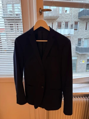 Klassisk svart kavaj Selected Homme - Stilren svart kavaj med två knappar framtill och smala slag. Kavajen har två fickor utan lock och en enkel, rak passform. Perfekt för en dressad look och funkar till många olika outfits. Materialet ser ut att vara en slät textil, troligen polyester eller ullblandning.