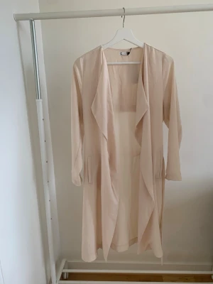 Beige lång trenchcoat med skärp - Stilren beige trenchcoat med öppen front och långa ärmar. Jackan har ett tunt, mjukt tyg och ett matchande skärp i midjan. Fickor på sidorna och klassisk slits baktill. Perfekt för lager på lager och en clean look.