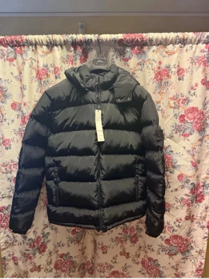 Svart dunjacka från Stone Island - Säljer en svart dunjacka från Stone Island med klassisk patch på ärmen. Jackan har huva, dragkedja framtill och två sidofickor med dragkedja. Puffig modell som håller dig varm under kalla vinterdagar. Materialet är glansigt och ger en modern look.
