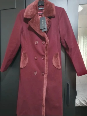 Vinröd lång kappa med fuskpäls - Stilren vinröd kappa från FengYunGu med dubbelknäppning och mjuka detaljer i fuskpäls vid krage och fickor. Kappan har en klassisk, lång siluett och är fodrad med matchande tyg. Perfekt för kalla dagar när du vill vara både varm och snygg.