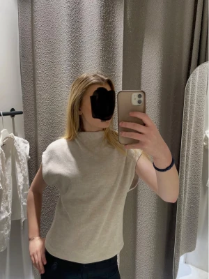 Beige ärmlös topp med hög krage - Stilren beige topp med hög krage och ärmlös design. Toppen har en rak passform och är tillverkad i ett mjukt material som ger en clean och modern look. Perfekt att matcha med jeans eller kjol för en enkel men snygg outfit.