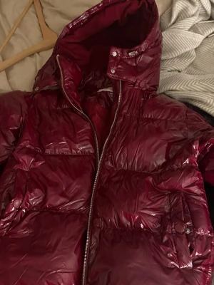 Vinröd glansig pufferjacka med huva -  Jag säljer otroligt fina och populära Nelly jacka som är otroligt skön och perfekt till kallas vinterdagar. Dock ser man att ena knappen till höger fickan är av (syns på bild) jackan är i still xxs