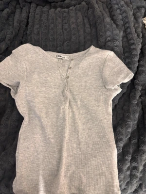 Ljusgrå ribbad topp från FB Sister - Säljer en ljusgrå ribbad topp från FB Sister i storlek S. Toppen har korta ärmar, rund halsringning och knappar framtill för en snygg detalj. Perfekt att matcha med jeans eller kjol för en enkel och stilren look.