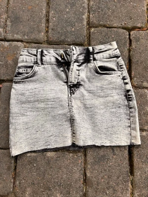 Grå kort jeanskjol med slitna detaljer - Säljer en grå kort jeanskjol från Bershka med slitna kanter och tvättad look. Svarta detaljer. Kjolen har fem fickor, bälteshällor och dragkedja framtill. Perfekt för dig som gillar en avslappnad och trendig stil.