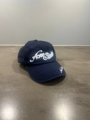 Mörkblå keps från Acne Studios - Snygg mörkblå keps från Acne Studios med broderad vit logga framtill och på skärmen. Justerbar baktill för perfekt passform. Tillverkad i 100% bomull vilket gör den både bekväm och lätt att bära!