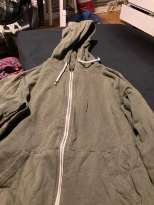 Olivgrön hoodie med dragkedja - Säljer en olivgrön hoodie med dragkedja och huva med vita snören. Hoodien har två fickor framtill och är tillverkad i mjukt bomullstyg. Perfekt för dig som gillar en avslappnad och enkel stil  Hämtas i Flocksta 3 i Västerås ändas  swhis