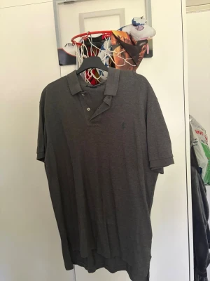 Mörkgrå pikétröja från Polo Ralph Lauren - Klassisk mörkgrå pikétröja från Polo Ralph Lauren med broderad logga på bröstet. Tröjan har krage, knappar framtill och korta ärmar. Perfekt till en avslappnad men ändå stilren look.