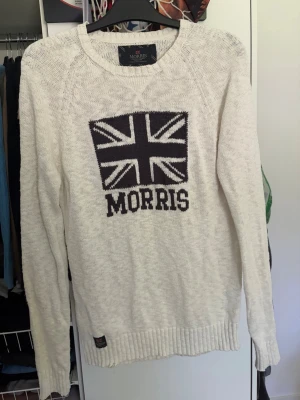 Vit stickad tröja från Morris - Vit stickad tröja från Morris med svart Union Jack-flagga och MORRIS-text på bröstet. Tröjan har rund halsringning, långa ärmar och ribbade muddar vid ärmslut och nederkant. Perfekt för dig som gillar klassisk och stilren design.