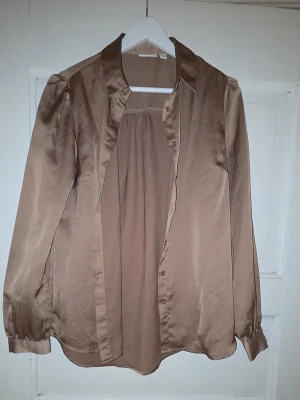 Beige satinliknande skjorta - Säljer en guldbeige skjorta i satinliknande material med klassisk krage och långa ärmar. Skjortan har knappar framtill och en avslappnad passform. Perfekt för dig som gillar stilrena och enkla plagg.
