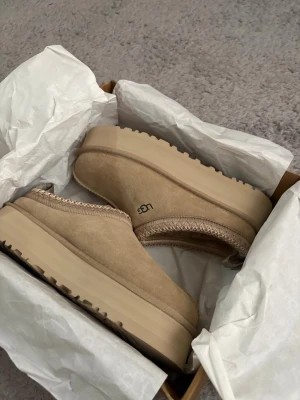 Beige UGG tofflor med platåsula - Säljer ett par beige UGG tofflor med kraftig platåsula och mjukt foder. Tofflorna är i mocka med dekorativ kant runt öppningen och har UGG-loggan på sidan. Perfekta för att hålla fötterna varma och bekväma hemma.