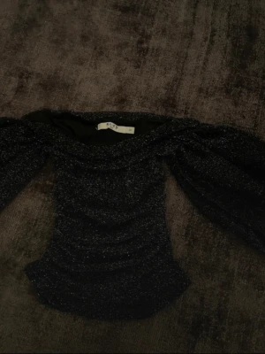Glittrig offshoulder topp från NA-KD - Säljer en svart glittrig offshoulder topp från NA-KD i storlek XS. Toppen har puffiga ärmar och är lite croppad med snygg rynkning framtill. Perfekt för dig som vill sticka ut med en trendig och festlig look till exempel på Nyår.