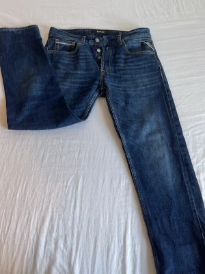 Mörkblå jeans från Replay - Snygga mörkblå jeans från Replay med raka ben och klassisk femficksmodell. Jeansen har knäppning med knappar och diskreta slitningar för en cool look. Materialet är denim i bomull med lite stretch för extra komfort.