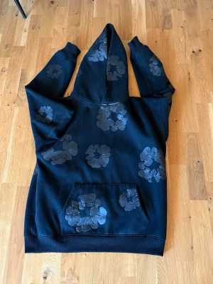 Denim tears hoodie - Säljer en svart hoodie från Denim Tears med unika blommönster i grått över hela plagget. Hoodien har en stor huva och en klassisk magficka framtill. Perfekt för dig som vill sticka ut med en streetwear-look.