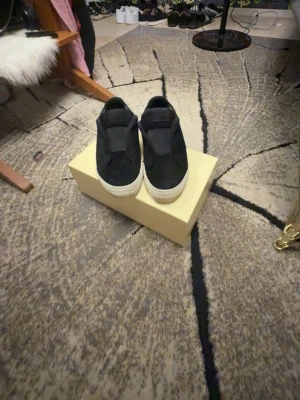Axel Arigato skor - Säljer ett par svarta slip-on sneakers från Axel Arigato. Skorna har en stilren design i mocka med vit sula och elastiska paneler på sidorna för enkel påtagning. Perfekta för dig som gillar minimalistisk och clean stil.