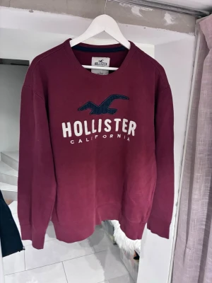 Vinröd sweatshirt från Hollister - M/L.  Vinröd sweatshirt från Hollister med stor logga och texten 'Hollister California' broderat på bröstet. Tröjan har rund halsringning, långa ärmar och mjukt material. Perfekt för en avslappnad och trendig look.