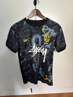 Svart Brasil x Stüssy t-shirt - Unik svart t-shirt med Brasil-logga och Stüssy-tryck framtill. T-shirten har korta ärmar och detaljerat grafiskt mönster i grått, gult och blått. Materialet känns som polyester, perfekt för en sportig och street-inspirerad look.