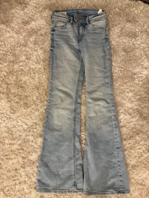 Ljusblå bootcut jeans - Säljer ett par ljusblå bootcut jeans med klassisk femficksdesign och hög midja. Jeansen har en snygg tvättad look och vida ben som ger en retrovibe. Perfekta för dig som gillar en avslappnad men trendig stil.