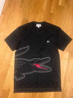 Svart Lacoste t-shirt med stor logga - Säljer en svart t-shirt från Lacoste med en stor krokodil-logga i grått och rött som sträcker sig från framsidan till baksidan. Klassisk rund hals och korta ärmar. Ikoniska lilla Lacoste-krokodilen på bröstet. Perfekt för dig som gillar streetwear och vill sticka ut.