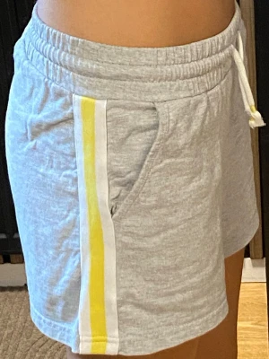 Grå shorts med gula ränder - Snygga grå shorts med vita och gula ränder längs sidorna. De har elastisk midja med snörning och två fickor framtill. Det är barn shorts, men sitter även som XS!
