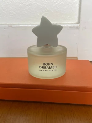 Born Dreamer Happy Place parfym - Born Dreamer Happy Place parfym i en frostad glasflaska med en unik stjärnformad vit kork. Flaskan har en rund form och texten är tryckt i svart på framsidan. Designen är modern och lekfull, perfekt för dig som gillar snygga detaljer på parfymhyllan.