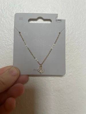 Guldigt halsband med bokstav M - Snyggt halsband i guldfärgad metall med ett litet hänge i form av ett M dekorerad med små glittrande stenar. Kedjan är tunn och elegant, perfekt för att ge en extra touch till din outfit. Passar dig som gillar detaljer och bling.
