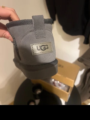 Gråa UGG tofflor i mocka - Säljer ett par gråa tofflor från UGG i mjuk mocka med klassisk UGG-logga. Helt nya med kartongen till