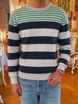 Vintage Randig stickad tröja J.Lindeberg - Snygg stickad tröja från J.Lindeberg med breda marinblå och ljusgrå ränder samt smalrandig grön och vit upptill. Rund halsringning och långa ärmar. Perfekt för dig som gillar klassisk men modern stil.