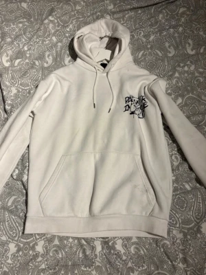Vit hoodie med tryck och björn - Säljer en vit hoodie med huva och känguruficka. På bröstet finns ett svart tryck med texten 'PARIS IN CRIME' och på ryggen ett stort tryck med en tecknad björn och ordet 'CRIME' upprepat. Perfekt för dig som gillar streetwear och unika prints.