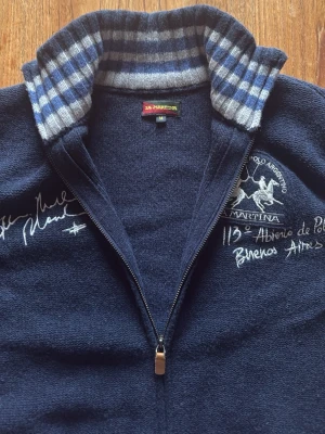 Marinblå ziptröja La Martina - Snygg marinblå ziptröja från La Martina med broderade detaljer och logga på bröstet. Tröjan har hög krage med rutigt foder i blått och vitt, samt ribbade muddar. Perfekt för dig som gillar sportig och stilren look.