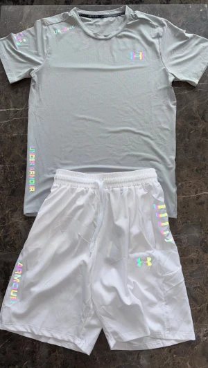 Grå/vit Under Armour set - Under Armour set med vita shorts och ljusgrå T-shirt. Setet har tryckta loggor och textdetaljer i både svart och holografisk färg i ljus/flash. Storlek M, helt ny med etiketterna kvar. Tveka inte att höra av vid frågor eller fler bilder. Kan säljas billigare vid snabb affär