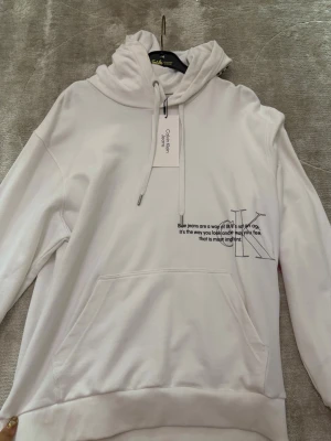 Vit hoodie med tryck från Calvin Klein - Snygg vit hoodie från Calvin Klein med stor huva och dragsko. Framtill finns ett tryck med text och en stor CK-logga. Hoodien har en klassisk känguruficka och ribbade muddar vid ärmslut och nederkant. Perfekt för en clean och modern look.