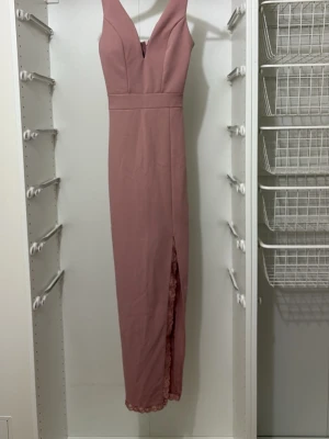 Lång rosa klänning med slits - Elegant långklänning i ljusrosa nyans med v-ringad urringning och breda axelband. Klänningen har en slits framtill och spetsdetalj längst ner. Perfekt för speciella tillfällen där du vill känna dig extra fin.