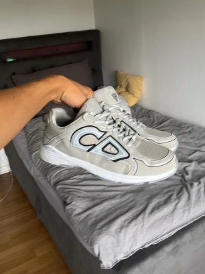 Gråa Dio sneakers med CD-logga - Snygga gråa sneakers från DlOR med stor CD-logga på sidan och detaljer i vitt och svart. Skorna har meshpaneler för extra ventilation och en vit sula som ger en sportig vibe. Perfekta för dig som vill ha något trendigt och exklusivt i garderoben.