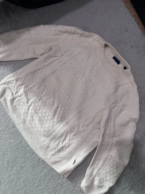 GANT Vit kabelstickad tröja - Säljer en krämvit kabelstickad tröja med rund hals och snygga detaljer i form av flätmönster och strukturstickning. Tröjan har långa ärmar och ribbade muddar vid ärmslut och nederkant. Perfekt för lager på lager under kyliga dagar.