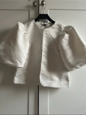 Bymalene liknande Vit blus med puffärmar  - Trendig vit blus med stora puffärmar och rak passform. Blusen har rund halsringning och är tillverkad i ett glansigt, strukturerat material som ger en lyxig känsla. Perfekt för dig som vill sticka ut med en statement-ärm.
