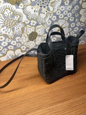 Svart flätad handväska från Lindex - Trendig svart handväska från Lindex med flätad struktur och dubbla handtag i konstläder. Väskan har även en lång axelrem och är fodrad med svart tyg invändigt. Perfekt för dig som gillar stilrena accessoarer med en twist. Nypris :349