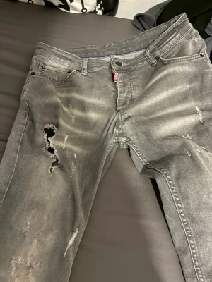 Grå slitna jeans med hål strlk w29 l 32 - Säljer ett par grå jeans med slitningar och hål på vänster lår. Jeansen har en klassisk femficksmodell och dragkedja. De har en tvättad look och är tillverkade i denim. Perfekta för en avslappnad och trendig stil.